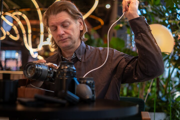 Man adjusting camera settings while seated at a table, surrounded by plants and soft lighting, showcasing the art of photography and equipment handling