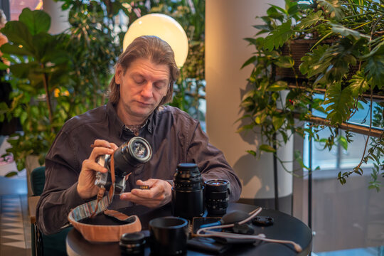 Man with long hair is adjusting camera lens while seated at a table surrounded by plants, showcasing photography equipment and tools in a creative workspace