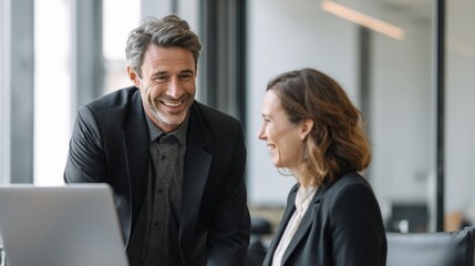 Man and woman in suits laughing together near a laptop in a bright office environment setting