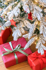 Beautiful presents, gift boxes wrapped in elegant red paper under green snowy festive Christmas tree with warm lights. Cozy winter holiday atmosphere perfect for seasonal celebrations and decor themes