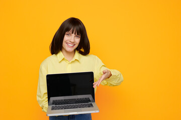 Fototapeta premium A cheerful woman stands against a vivid orange backdrop, proudly presenting a laptop. This image conveys online learning, remote work, and modern technology with approachable energy.