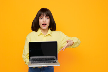 Naklejka premium A surprised young woman presents an open laptop toward the camera, standing against a bright orange backdrop. She wears a pale yellow shirt, creating a cheerful, modern tech vibe ideal for digital