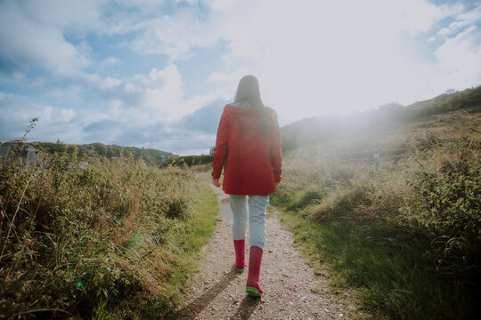 A woman in a red jacket and pink rain boots walks away on a sun-drenched, overgrown dirt path through a grassy coastal landscape, under a bright, cloudy sky. Sun flare creates a vibrant, warm mood. - Powered by Adobe