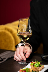A person's hand holds a glass of white wine at a fine dining table, with gourmet appetizers in the foreground. The image features a close-up, shallow depth of field, and warm, elegant lighting