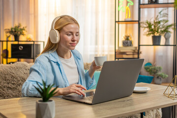 Young woman manages tasks at home, typing on laptop with wireless headphones while enjoying warm...
