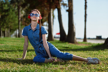 Fototapeta premium Young woman in blue jumpsuit relaxing on grass with sunglasses under bright sunny sky near palm trees, enjoying warm weather and casual outdoor style.