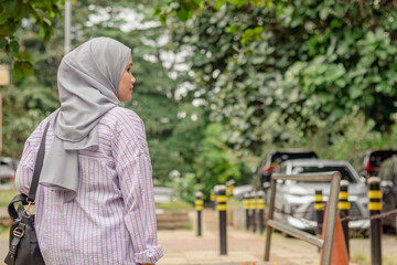 A young Asian Muslim woman carrying a bag on her shoulder while walking towards an outdoor parking...