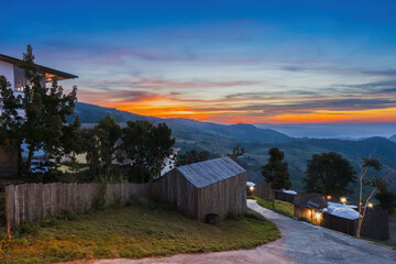 Sunset over rural hillside with wooden houses, trees, and winding road creating peaceful and scenic countryside atmosphere