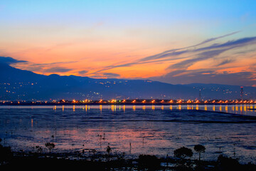 Obraz premium Stunning twilight scenery over Wugu Wetland in New Taipei, Taiwan. Mountains silhouetted against golden sky, city lights reflecting on calm waters.