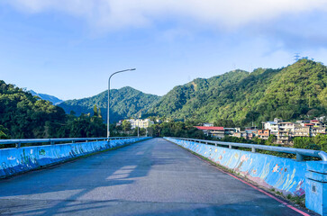Empty bridge with vibrant blue ocean mural on barriers leading to mountain village in Wulai, New Taipei City, Taiwan. Early morning light and lush green hills.