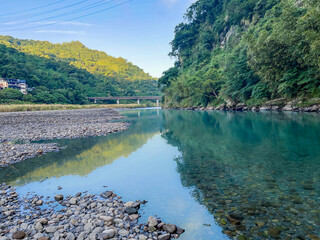 The turquoise water of the river flows past a rocky gravel bank in Wulai, New Taipei City, Taiwan. Lush green mountain slopes frame the clear stream with a distant bridge visible on a sunny morning.