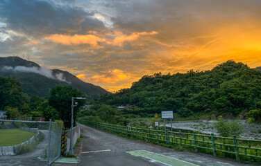 Vibrant sunrise with fiery orange clouds above the misty green mountains of Wulai, New Taipei City, Taiwan. A quiet riverside road leads into the lush valley at dawn.