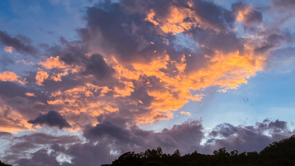 Spectacular dawn cloudscape in Wulai, New Taipei City, Taiwan. Vibrant orange and golden clouds contrast with blue sky above silhouetted mountain treeline.