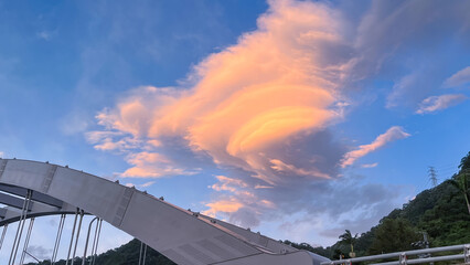 Unique lenticular cloud formation illuminated with warm orange light above the modern bridge structure in Wulai New Taipei City Taiwan during dawn.