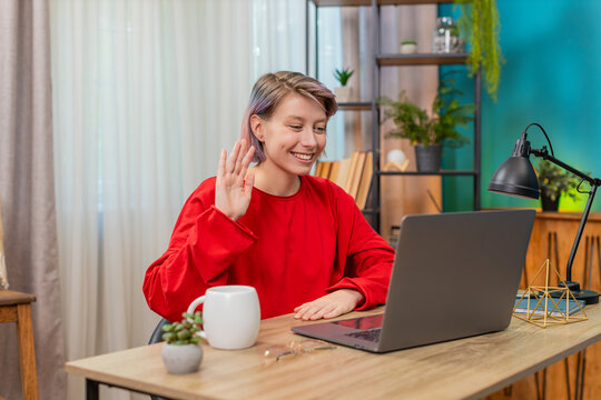 Young woman making online negotiation on laptop sitting at home table, smiling and waving hand cheerfully. Caucasian girl demonstrating friendly, satisfied attitude, enjoying video call communication - Powered by Adobe