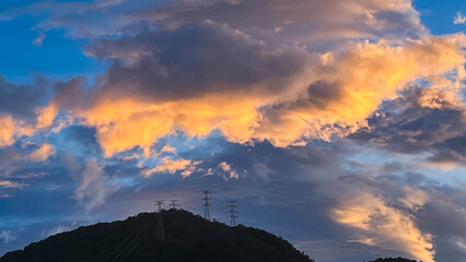 Stunning sky over Wulai New Taipei City Taiwan at dawn or dusk. Dramatic orange and gray clouds hover above power transmission towers on a forested mountain.