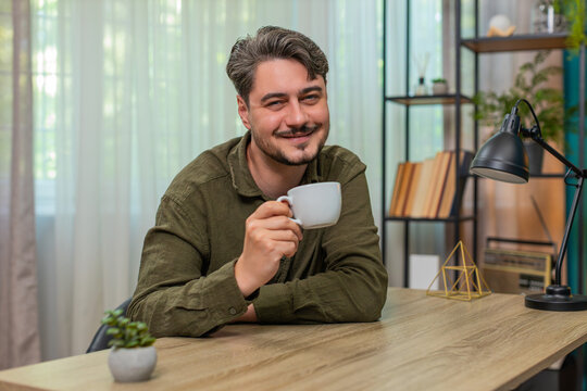 Middle-aged man at home holds mug sipping coffee enjoying tranquil relaxation moment feeling peace. Young adult guy at table breathes slowly calm mood contentment resting before continuing tasks today