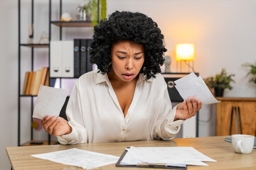 African American businesswoman at home office views bills on invoices feeling stressed about debt and taxes. Freelancer girl at table holds receipts worried by fees budget gap cuts to avoid bankruptcy