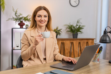 Middle-aged businesswoman at home office works on laptop drinks coffee takes sip enjoys aroma smiles. Freelancer girl at table warms hands on cup gets energy boosts mood continues tasks types steady.