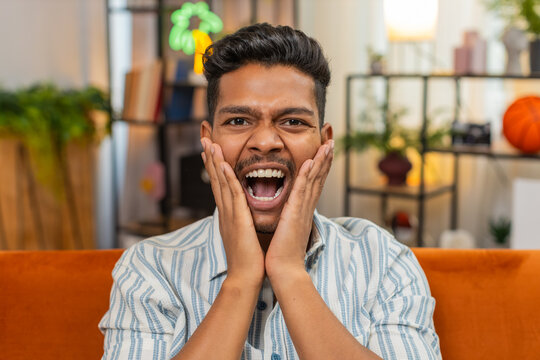 Indian man at home grabs cheeks and says wow after reading positive news on smartphone and smiles. Arabian guy on sofa feels surprise opens mouth raises eyebrows claps hands and shows joyful reaction.