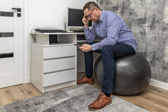 Male office worker in blue shirt sitting on orthopedic ball in front of computer, office workers back problems