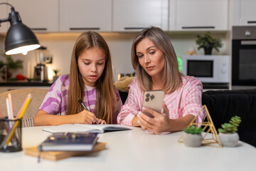 Mother and daughter watch online math lesson on smartphone mother explains example child writes notes in notebook listens to tip studies together. Woman with teenager girl sitting at table at evening.