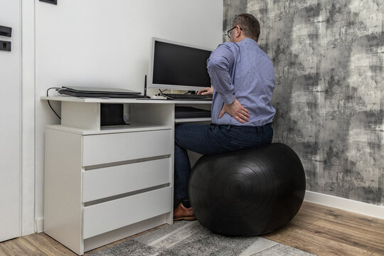 Male office worker in blue shirt sitting on orthopedic ball in front of computer, office workers back problems