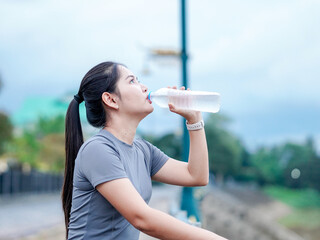 Asian fitness woman drinking water bottle after runners workout at park motivation focus new year, cardio, self care.Happy Female light exercise routine. Healthy lifestyle wellness. Healthy choice