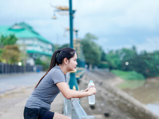 Asian fitness woman drinking water bottle after runners workout at park motivation focus new year, cardio, self care.Happy Female light exercise routine. Healthy lifestyle wellness. Healthy choice