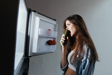 Young woman in casual attire biting cucumber in front of open fridge, showcasing healthy eating...