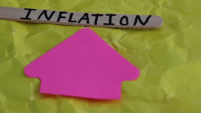 Un primer plano de una paleta de madera con la palabra de inflaci&oacute;n y una flecha de color rosado con movimiento en un fondo amarillo. Pol&iacute;tica econ&oacute;mica 