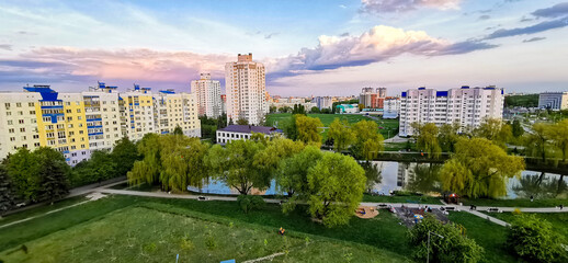 Naklejka premium High-angle view of modern residential district with apartment buildings surrounding a green city park and pond at sunset. Blend of urban planning and urban nature.