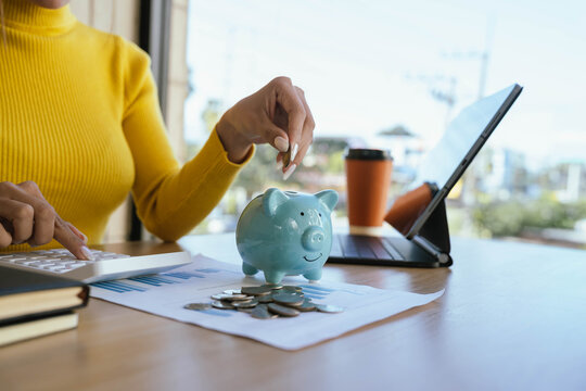 A person in a yellow sweater places a coin into a blue piggy bank on a desk with charts, coins, a laptop, and a coffee cup, symbolizing saving and financial planning. - Powered by Adobe