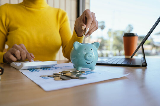 A person in a yellow sweater places a coin into a blue piggy bank on a desk with charts, coins, a laptop, and a coffee cup, symbolizing saving and financial planning.
