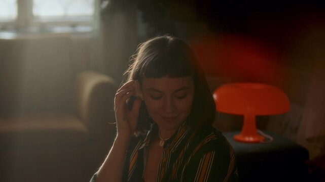 Young woman with bob haircut holding seashells to her ears, listening to them and smiling in cozy warmly lit living room with retro lamp