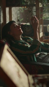 Young dreamy woman leaning back in armchair, holding shell to ear and examining it as relaxing in cozy room filled with plants and warm light. Vertical clip