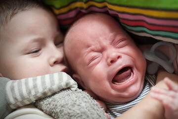 A toddler leans close to a crying baby, offering comfort in a cozy environment. The siblings share a moment under colorful blankets, showcasing tenderness and connection during playtime.