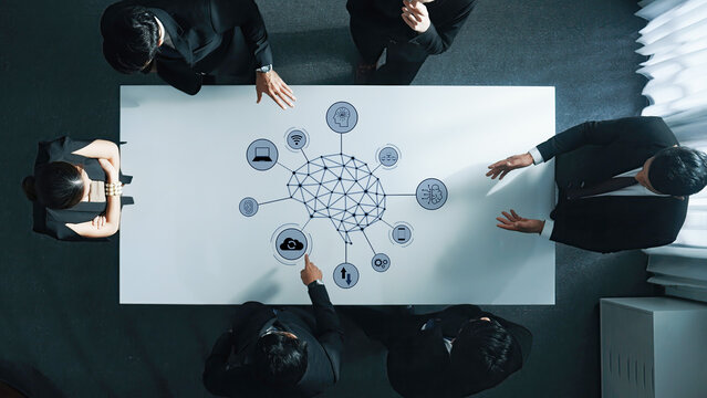 A diverse group of business professionals engages in a strategic meeting around a modern conference table, focusing on technology graphics that illustrate network connections. Trope