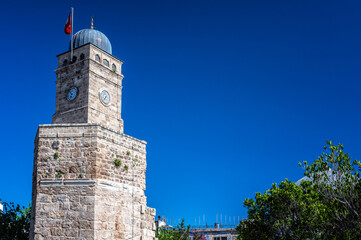 The historic clock tower of Antalya, Turkey, topped with a Turkish flag under a deep blue sky. A notable city monument and symbol of heritage.