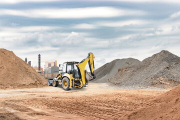 Obraz premium A yellow backhoe works on a construction site, digging through mounds of dirt and gravel with industrial buildings in the background on a cloudy day