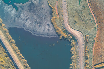 Abstract drone view capturing the complex, double-track boundary dividing dark, contaminated industrial lake from lush green vegetation and a brown field. Severity of the ecological crisis.