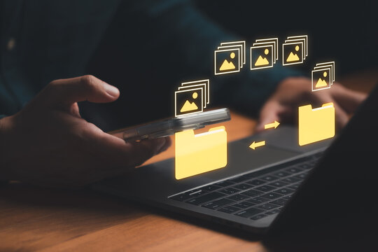 Man holding mobile phone with digital image file icon and a folder graphic between the phone and a laptop, representing photo transfer, cloud backup, data syncing, and cross device file management.