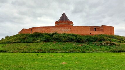 Red brick fortress castle Lindau with round tower on grassy hill beneath cloudy gray sky