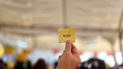 Left hand holding queue ticket number N100 printed in black ink on yellow paper with blur...