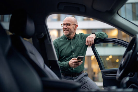 Smiling mature businessman with glasses holding smartphone leaning against open car door in urban city