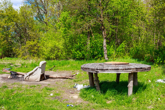 Rustic wooden table and torn armchair mark a messy outdoor hangout spot amidst vibrant spring greenery