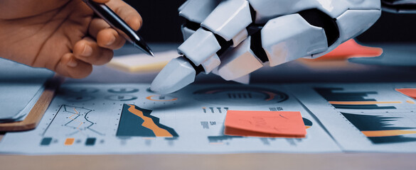 A human hand and a robotic hand work together on business charts, analyzing data trends and showcasing the fusion of technology and human creativity in the workplace.Noogenesis