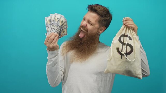 Man holding moneybag and fanning hundred dollar bills with playful grin in studio against teal backdrop; cheerfulness.