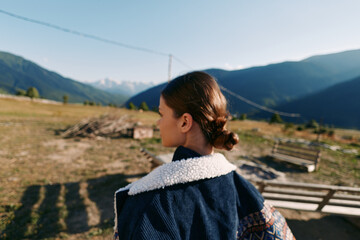 Fototapeta premium Girl portrait outdoors in mountains countryside, back view of woman wearing jacket, rural landscape with fields and wooden fence, calm travel scene and warm natural summer sunlight.