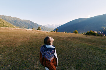 Fototapeta premium Woman in coat stands in a meadow field looking up at sky and distant mountains, serene landscape and travel concept with open space, solitude and nature scenery.
